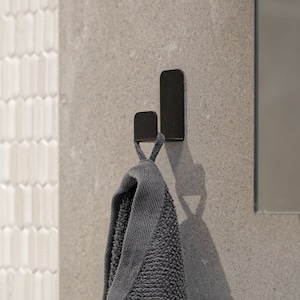 May include: A matte black metal wall hook with a dual-prong design, featuring a shorter front hook and a taller back hook. A dark gray textured cotton towel is hanging from the front prong of the hook, which is mounted on a flat stone surface.