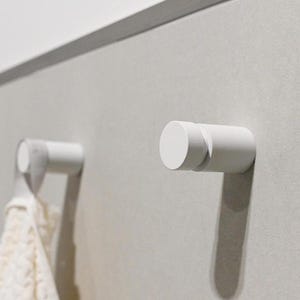 May include: Two cylindrical white wall hooks mounted on a flat surface. One hook is empty, while the other hook holds a white textured fabric item with a loop. The hooks feature a smooth, matte finish and a simple, uniform design for hanging items.