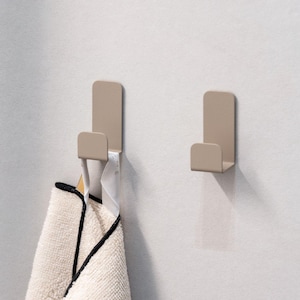 Op de afbeelding: Twee beige metalen handdoekhaken aan een witte betegelde muur. Eén haak houdt een crèmekleurige handdoek met zwarte rand vast. De haken zijn eenvoudig en modern, voor badkamer of keuken.