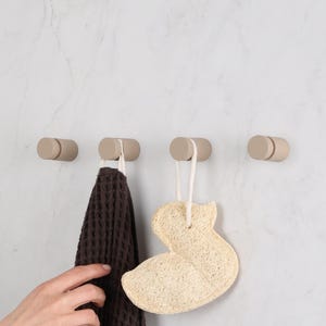 Può includere: Quattro ganci da parete cilindrici color beige montati in una fila orizzontale. Un asciugamano marrone scuro con trama a nido d'ape pende dal secondo gancio, e una spugna di luffa color cuoio a forma di papera pende dal terzo gancio.
