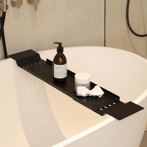 Könnte beinhalten: Eine mattschwarze Badewannenablage aus Metall mit perforierter Oberfläche, die auf den Rändern einer weißen freistehenden Badewanne liegt. Die Ablage enthält eine braune Glas-Pumpeflasche mit weißem Etikett, einen weißen Tiegel und eine weiße Tube.