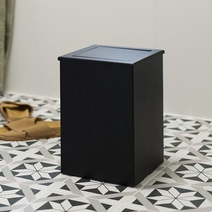 Puede incluir: Un cubo de basura rectangular de color negro mate con tapa plana y bordes limpios y definidos. El recipiente presenta un acabado sólido y opaco con un diseño sencillo y funcional, colocado sobre una superficie con un patrón de baldosas.