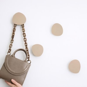 Pode incluir: Uma bolsa de mão cor taupe com uma alça de corrente está pendurada em um gancho de parede correspondente. Vários outros ganchos em forma de seixo são montados na parede branca. A bolsa tem um topo curvo e uma alça pequena.