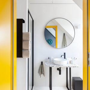 May include: A bathroom interior featuring a white vessel sink on a speckled terrazzo countertop with black metal legs. A round mirror hangs above the sink, accompanied by a black faucet, a black towel rack with rolled towels, and a black trash bin.