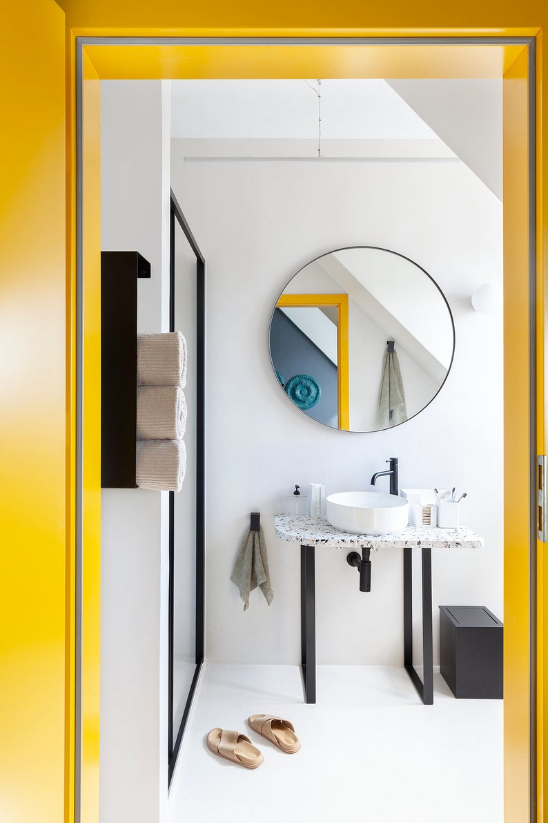 May include: A bathroom interior featuring a white vessel sink on a speckled terrazzo countertop with black metal legs. A round mirror hangs above the sink, accompanied by a black faucet, a black towel rack with rolled towels, and a black trash bin.