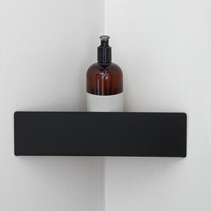 May include: A matte black corner shelf mounted on a white wall, featuring a rectangular front panel. A brown amber-colored plastic bottle with a black pump dispenser and a plain white label sits on the surface of the shelf.