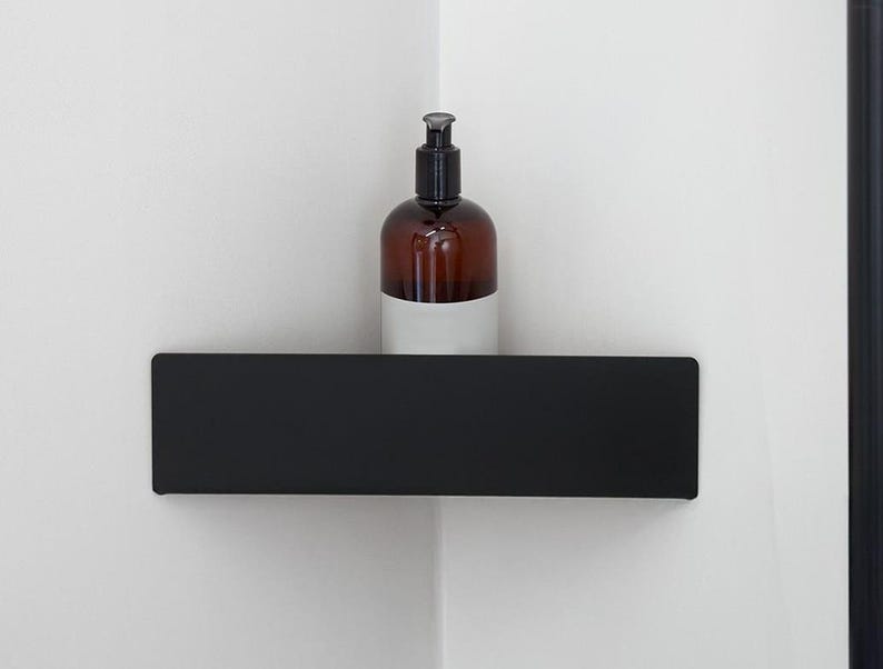 May include: A matte black corner shelf mounted on a white wall, featuring a rectangular front panel. A brown amber-colored plastic bottle with a black pump dispenser and a plain white label sits on the surface of the shelf.