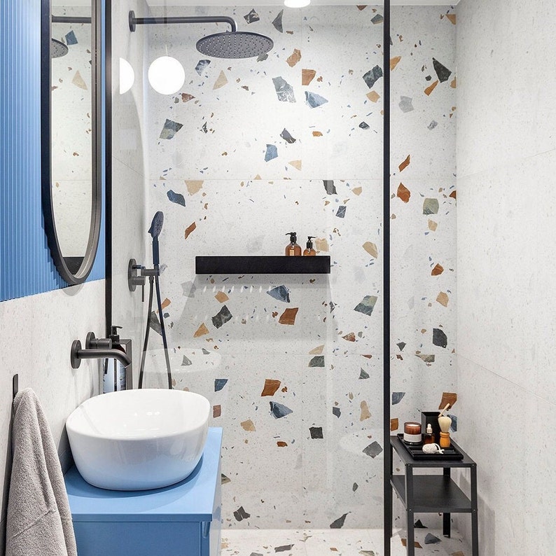 May include: A modern bathroom with a walk-in shower featuring a white tiled wall with brown and blue terrazzo accents. The shower has a black shower head and a black shower rod. A black shelf is mounted on the wall above the shower head. A blue vanity with a white sink is in the foreground.
