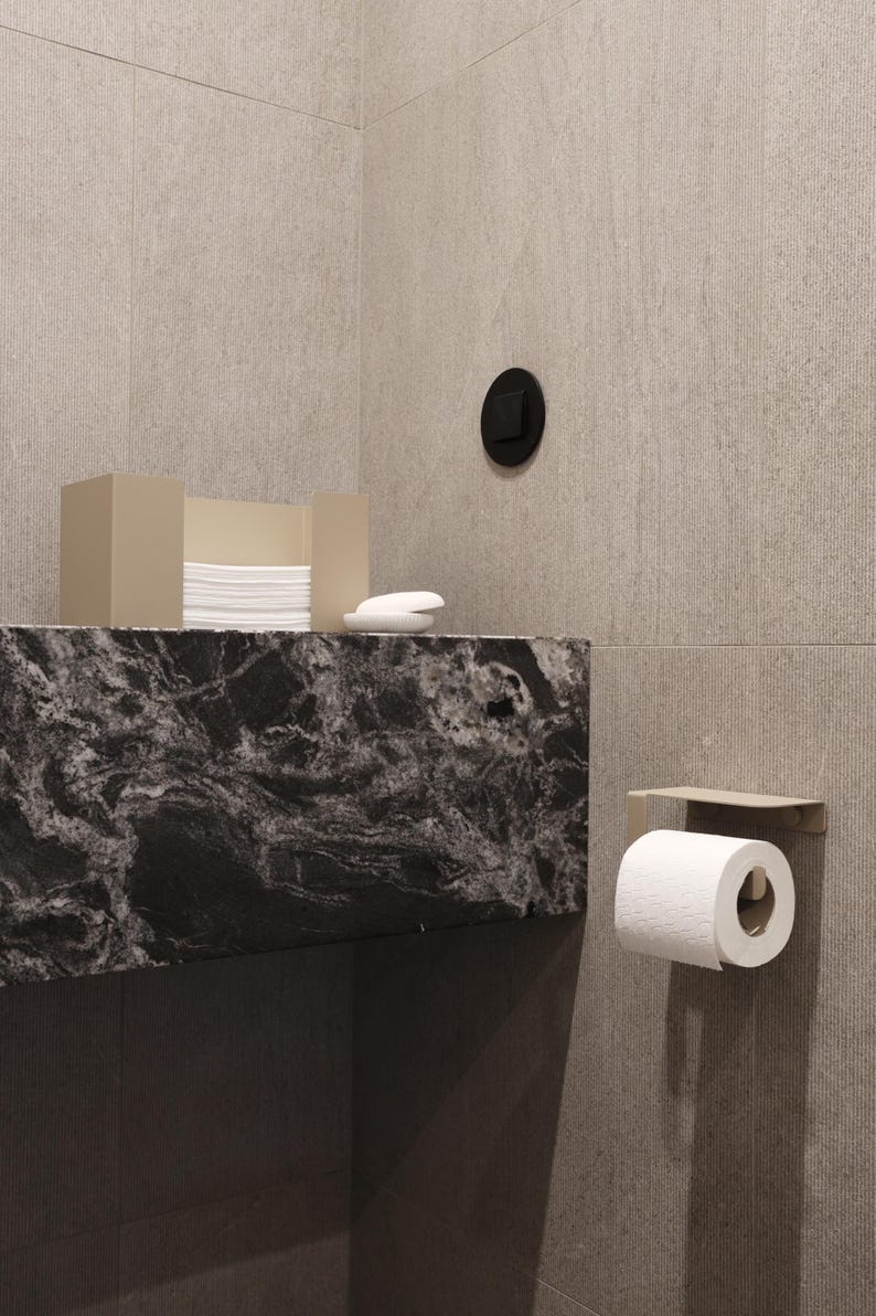 Puede incluir: Una escena de ba&ntilde;o moderno con una encimera de m&aacute;rmol negro y gris, una caja de pa&ntilde;uelos y jab&oacute;n. Un rollo de papel higi&eacute;nico est&aacute; montado en la pared. Las paredes son de color beige claro.