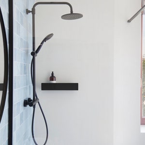 May include: A black shower head and handheld shower with a black shelf and a bottle of soap. The shower is in a bathroom with a white wall and a window with a view of a city.