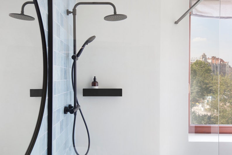 May include: A black shower head and handheld shower with a black shelf and a bottle of soap. The shower is in a bathroom with a white wall and a window with a view of a city.