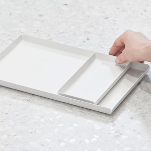 May include: A white metal tray with a smaller white metal tray inside. The trays are nested together and have a modern, minimalist design.