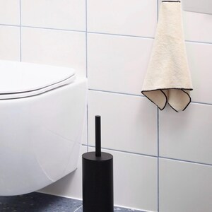 May include: A matte black cylindrical toilet brush holder with a matching handle, positioned next to a white toilet. A cream-colored hand towel with a thin black border hangs from a dark red metal hook mounted on the white tiled wall.