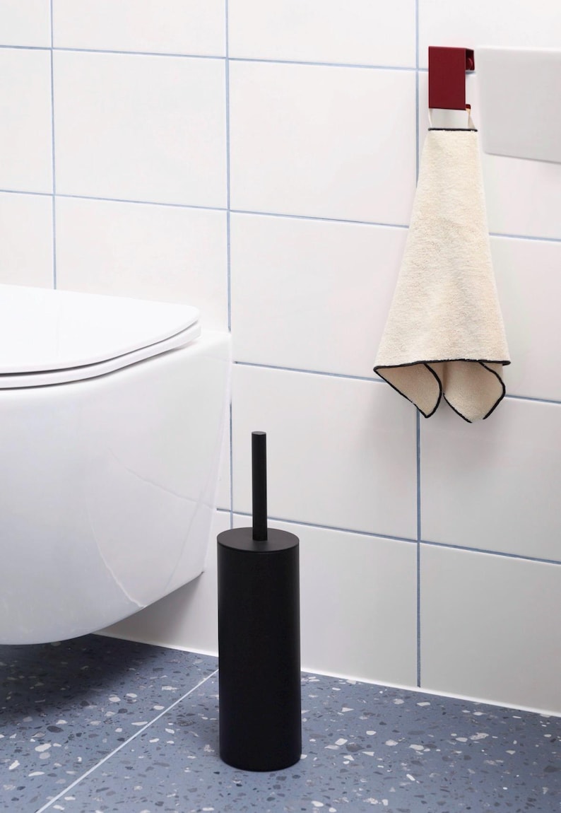 May include: A matte black cylindrical toilet brush holder with a matching handle, positioned next to a white toilet. A cream-colored hand towel with a thin black border hangs from a dark red metal hook mounted on the white tiled wall.