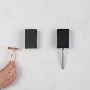 May include: A person holds a rose gold safety razor, positioned near two black razor holders. One holder is empty, and the other contains a silver razor. The items are set against a white marble backdrop.