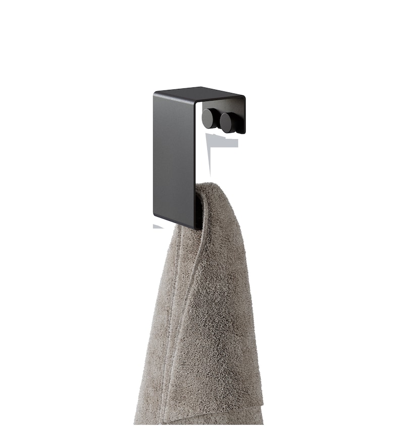 May include: A matte black metal over-the-door hook featuring a flat rectangular front panel and two small circular knobs on the inner side, with a folded light brown textured towel draped over the lower section of the hook structure.