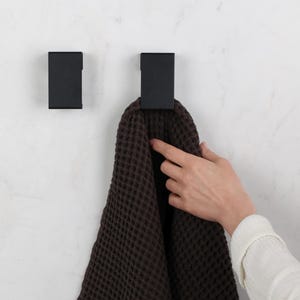 May include: Two rectangular matte black wall hooks mounted on a white surface. One hook holds a dark brown waffle-textured towel. The hooks feature a clean, geometric design with a flat front face and a simple, functional structure for hanging items.