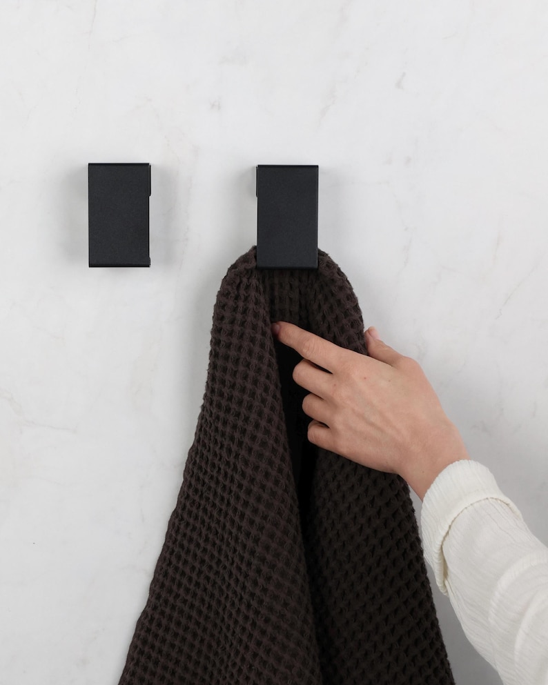 May include: Two rectangular matte black wall hooks mounted on a white surface. One hook holds a dark brown waffle-textured towel. The hooks feature a clean, geometric design with a flat front face and a simple, functional structure for hanging items.