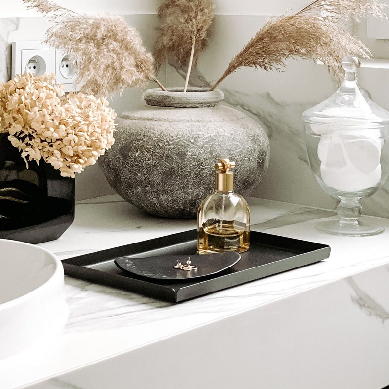 May include: A black tray with a small gold dish and a glass bottle with a gold top on a white marble countertop. A vase with dried pampas grass and a glass jar with cotton balls are also visible.