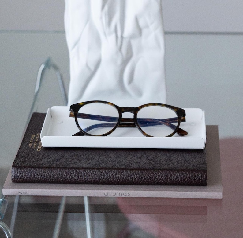 May include: A white rectangular tray with a pair of tortoiseshell eyeglasses on top. The tray is sitting on a brown leather-bound journal with the word "aromas" printed on the front.