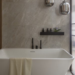 May include: A white bathtub with a towel draped over the edge. The bathtub is in a bathroom with gray tile walls and a black shelf with bottles and a plant on it.