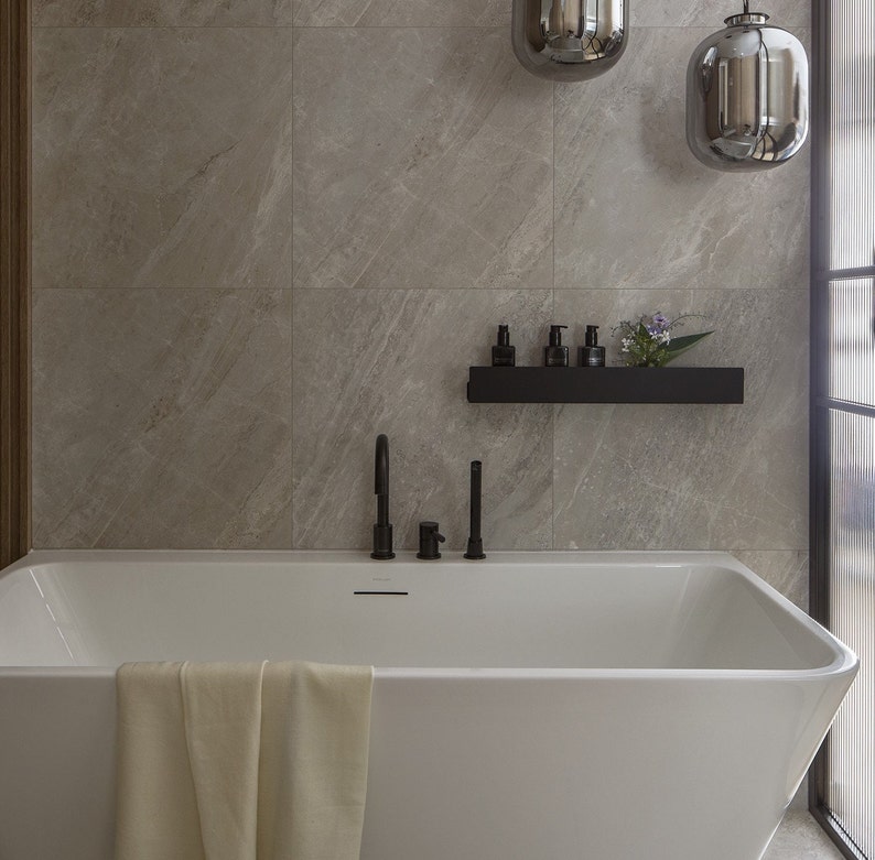 May include: A white bathtub with a towel draped over the edge. The bathtub is in a bathroom with gray tile walls and a black shelf with bottles and a plant on it.