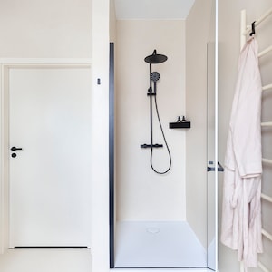 May include: A bathroom interior featuring a walk-in shower with a matte black rainfall showerhead, handheld sprayer, and matching shelf. A white door with a black handle is adjacent to the shower, and a beige robe hangs on a white towel rack.