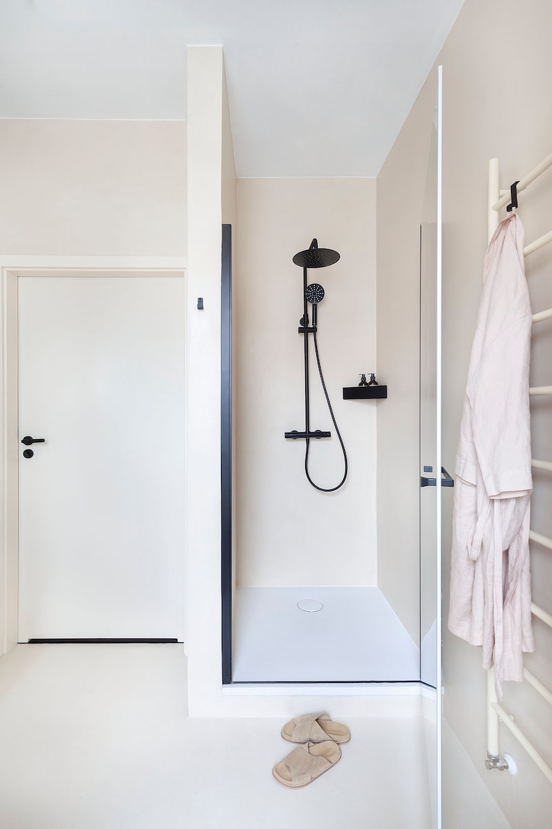 May include: A bathroom interior featuring a walk-in shower with a matte black rainfall showerhead, handheld sprayer, and matching shelf. A white door with a black handle is adjacent to the shower, and a beige robe hangs on a white towel rack.