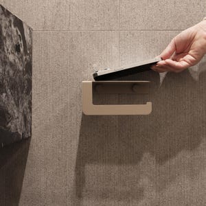 Puede incluir: Un portarrollos de papel higi&eacute;nico beige montado en una pared texturizada de color marr&oacute;n claro. Una mano coloca un tel&eacute;fono inteligente en el soporte. El soporte tiene un dise&ntilde;o simple y moderno.