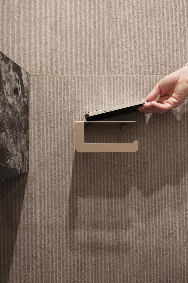 Puede incluir: Un portarrollos de papel higi&eacute;nico beige montado en una pared texturizada de color marr&oacute;n claro. Una mano coloca un tel&eacute;fono inteligente en el soporte. El soporte tiene un dise&ntilde;o simple y moderno.