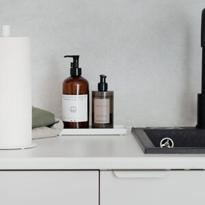 May include: White countertop with a white paper towel holder, a brown bottle of hand soap with the text "Savon Liquide de Marseille", a brown bottle of body wash with the text "Zara Home", and a white ceramic sink with a black faucet.