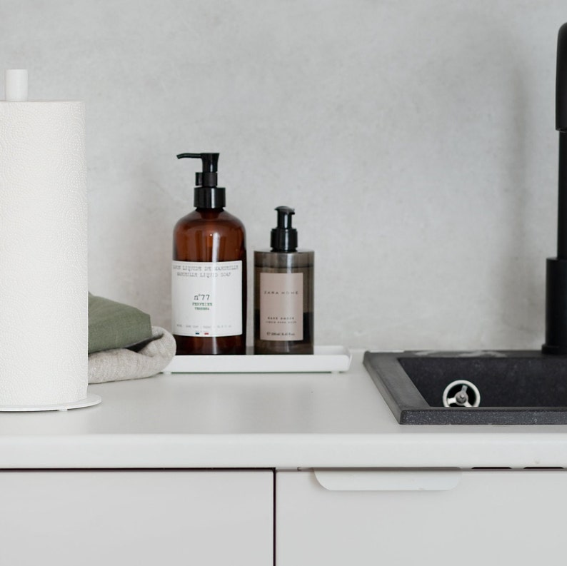 May include: White countertop with a white paper towel holder, a brown bottle of hand soap with the text "Savon Liquide de Marseille", a brown bottle of body wash with the text "Zara Home", and a white ceramic sink with a black faucet.