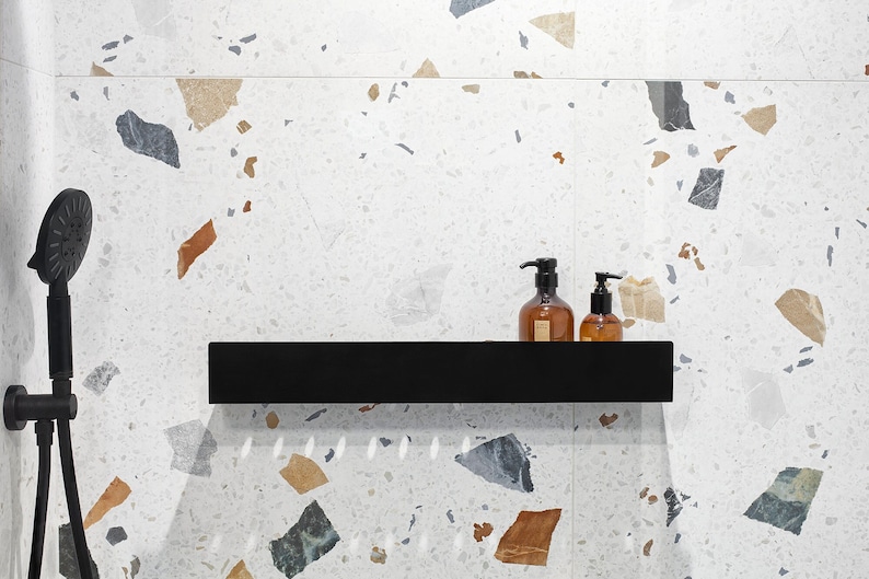 May include: A black shower head and arm are mounted on a white tiled wall with a terrazzo pattern. A black shelf is mounted on the wall with two amber glass bottles with pumps on top.