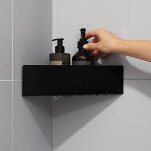 May include: A matte black corner shower shelf mounted against light gray tiled walls. The shelf holds two dark amber glass bottles with black pump dispensers, each featuring a label with printed text. The shelf surface has visible water droplets.