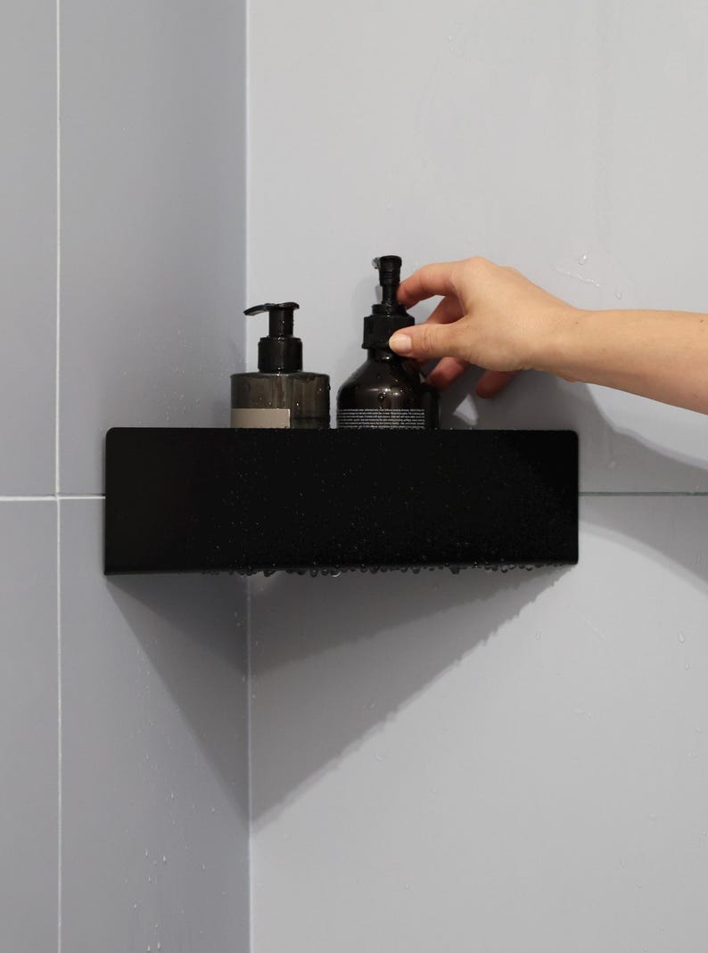 May include: A matte black corner shower shelf mounted against light gray tiled walls. The shelf holds two dark amber glass bottles with black pump dispensers, each featuring a label with printed text. The shelf surface has visible water droplets.