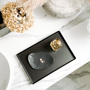 May include: A black tray with a small black dish and a clear glass bottle with a gold lid. The tray is on a white marble surface.
