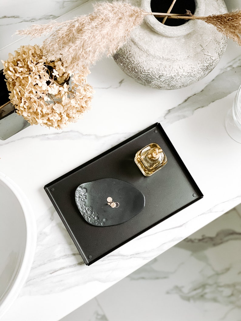 May include: A black tray with a small black dish and a clear glass bottle with a gold lid. The tray is on a white marble surface.