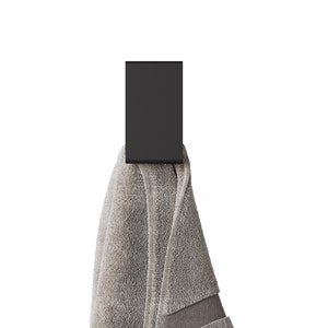 May include: A matte black rectangular wall hook holding a folded light gray towel. The hook features a flat, vertical design with a curved bottom edge, while the towel displays a soft, textured fabric surface with a visible hemmed border detail.