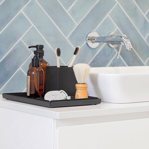 May include: A black tray with two brown glass bottles with pumps, a black toothbrush holder with two toothbrushes, a wooden shaving brush, and a gray pumice stone. The tray is on a white countertop in front of a blue tiled wall.