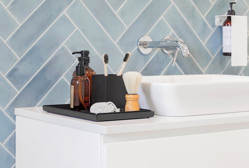 May include: A black tray with two brown glass bottles with pumps, a black toothbrush holder with two toothbrushes, a wooden shaving brush, and a gray pumice stone. The tray is on a white countertop in front of a blue tiled wall.