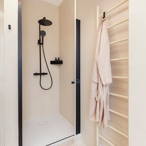 May include: A walk-in shower with a matte black rainfall showerhead, handheld sprayer, and black frame. A beige robe hangs on a cream-colored towel warmer rack. A pair of beige slide sandals rests on the floor near the shower entrance.