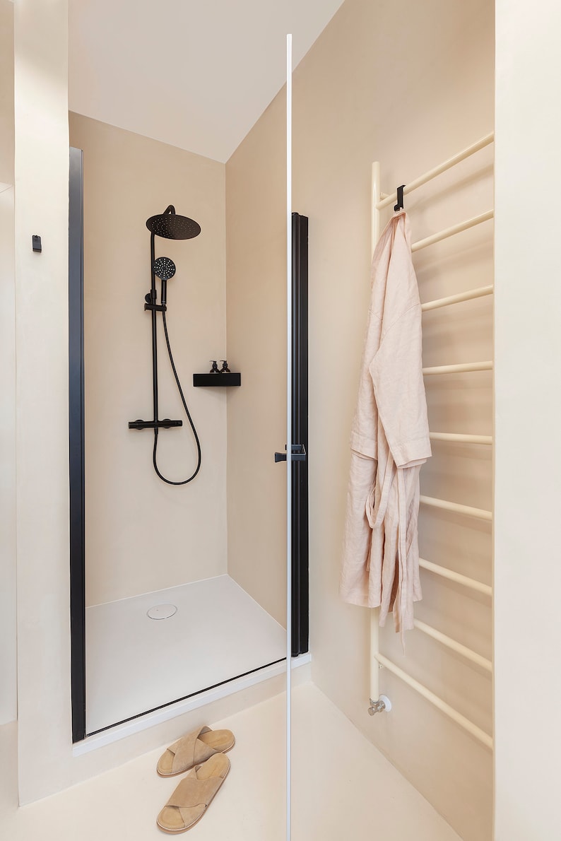May include: A walk-in shower with a matte black rainfall showerhead, handheld sprayer, and black frame. A beige robe hangs on a cream-colored towel warmer rack. A pair of beige slide sandals rests on the floor near the shower entrance.