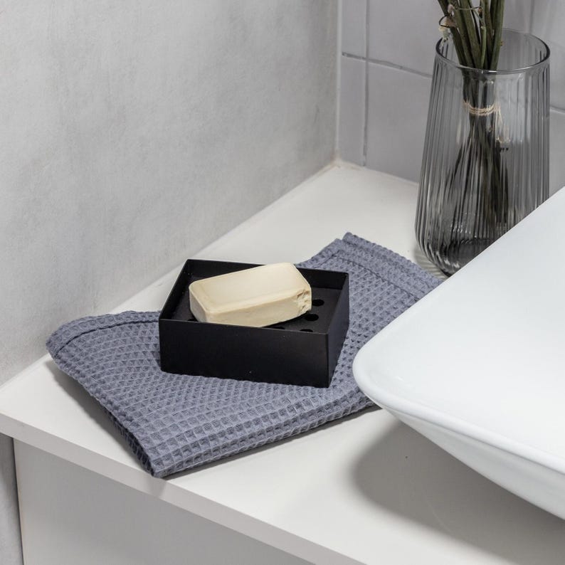 Może przedstawiać: Biała ceramiczna umywalka łazienkowa z szarym ręcznikiem wafelkowym i czarną mydelniczką z kostką mydła na białym blacie.