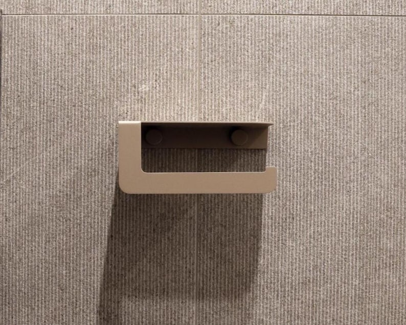 Puede incluir: Un portarrollos de papel higi&eacute;nico moderno y minimalista de metal cepillado. El soporte est&aacute; montado en una pared de azulejos texturizados de color gris claro. El dise&ntilde;o presenta un brazo en forma de L.