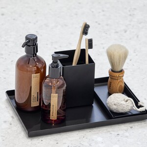 May include: A black metal tray with two amber glass bottles with pumps, a black container with two wooden toothbrushes, a wooden shaving brush with white bristles, and a white pumice stone.