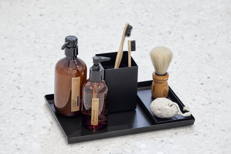 May include: A black metal tray with two amber glass bottles with pumps, a black container with two wooden toothbrushes, a wooden shaving brush with white bristles, and a white pumice stone.