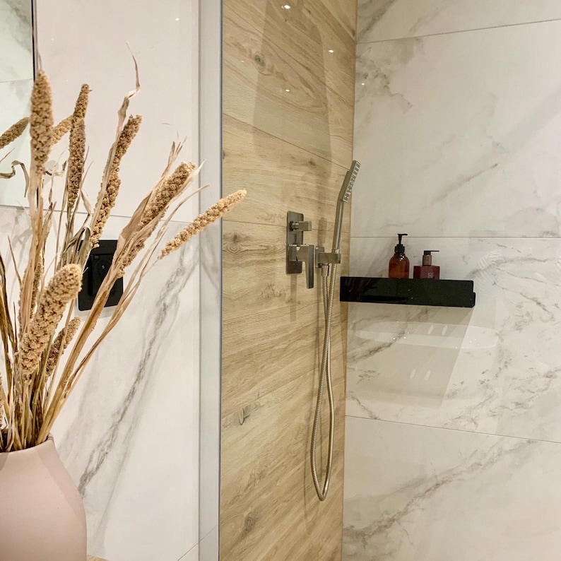 May include: A modern bathroom shower with a wood-look tile wall and white marble tile walls. The shower head is silver and has a handheld shower wand. A black shelf with two bottles of soap is mounted on the wall.