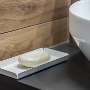 May include: A white ceramic soap dish with a white bar of soap inside. The dish is sitting on a dark gray countertop.