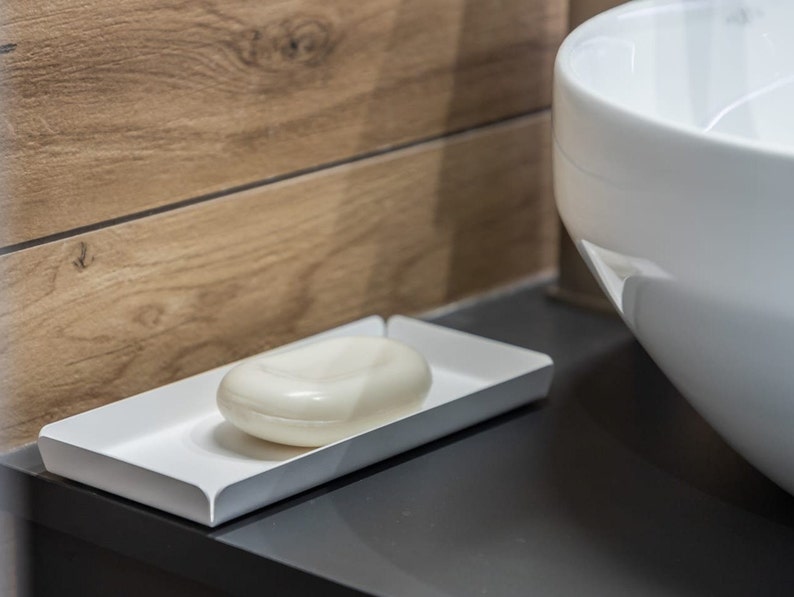 May include: A white ceramic soap dish with a white bar of soap inside. The dish is sitting on a dark gray countertop.