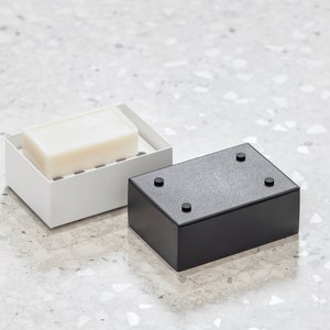 May include: A white soap dish with a bar of soap inside, next to a black soap dish with four rubber feet on the bottom.